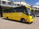 A yellow school bus is parked in front of a grey building with a corrugated metal roof. The bus has the name and contact information of a high school printed on its side, along with the school's motto. A few individuals are standing near the bus, and a white vehicle is visible in the background.