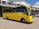 A yellow school bus is parked in front of a grey building with a corrugated metal roof. The bus has the name and contact information of a high school printed on its side, along with the school's motto. A few individuals are standing near the bus, and a white vehicle is visible in the background.