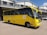 A yellow school bus is parked in front of a grey building with a corrugated metal roof. The bus has the name and contact information of a high school printed on its side, along with the school's motto. A few individuals are standing near the bus, and a white vehicle is visible in the background.
