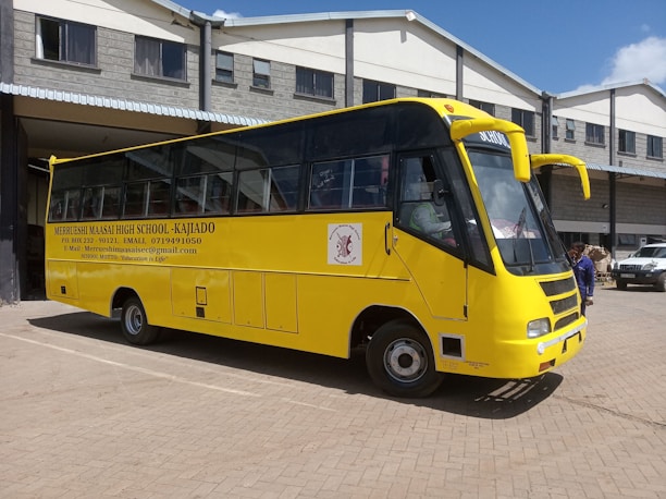 A yellow school bus is parked in front of a grey building with a corrugated metal roof. The bus has the name and contact information of a high school printed on its side, along with the school's motto. A few individuals are standing near the bus, and a white vehicle is visible in the background.