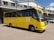 A yellow school bus is parked in front of a grey building with a corrugated metal roof. The bus has the name and contact information of a high school printed on its side, along with the school's motto. A few individuals are standing near the bus, and a white vehicle is visible in the background.