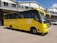 A yellow school bus is parked in front of a grey building with a corrugated metal roof. The bus has the name and contact information of a high school printed on its side, along with the school's motto. A few individuals are standing near the bus, and a white vehicle is visible in the background.