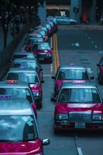 A row of red taxis is lined up along a city street, each with the word 'TAXI' illuminated on top. The vehicles are parked closely on the left side of the road with their roofs reflecting the surrounding cityscape. The background shows a few buildings and traffic lights.