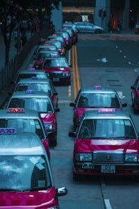 A row of red taxis is lined up along a city street, each with the word 'TAXI' illuminated on top. The vehicles are parked closely on the left side of the road with their roofs reflecting the surrounding cityscape. The background shows a few buildings and traffic lights.