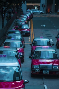 A row of red taxis is lined up along a city street, each with the word 'TAXI' illuminated on top. The vehicles are parked closely on the left side of the road with their roofs reflecting the surrounding cityscape. The background shows a few buildings and traffic lights.