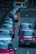 A row of red taxis is lined up along a city street, each with the word 'TAXI' illuminated on top. The vehicles are parked closely on the left side of the road with their roofs reflecting the surrounding cityscape. The background shows a few buildings and traffic lights.