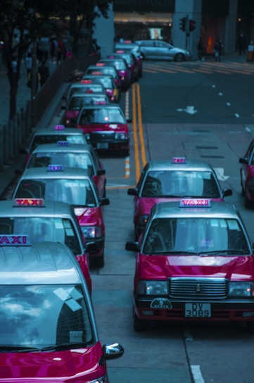 A row of red taxis is lined up along a city street, each with the word 'TAXI' illuminated on top. The vehicles are parked closely on the left side of the road with their roofs reflecting the surrounding cityscape. The background shows a few buildings and traffic lights.