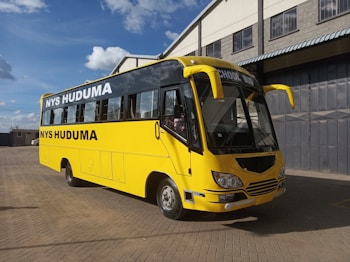 A bright yellow bus with black lettering indicating 'NYS Huduma' stands on a paved surface. The words 'School Bus' are displayed at the front. The bus is parked in an open area with a large industrial building nearby, under a blue sky with scattered clouds.