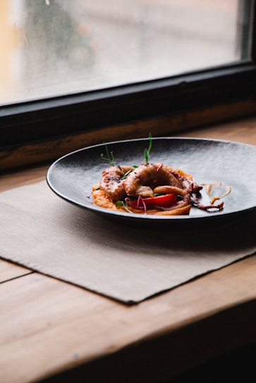 A plate of gourmet food is placed near a window on a wooden table. The dish consists of what appears to be a piece of grilled octopus garnished with herbs and vegetables, presented on a dark-colored plate with a textured design.