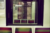 A train interior with retro-style seats in a pattern of vibrant colors. The seats are empty, and there are subway maps and station signs visible through the window, casting reflections.
