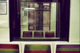 A train interior with retro-style seats in a pattern of vibrant colors. The seats are empty, and there are subway maps and station signs visible through the window, casting reflections.