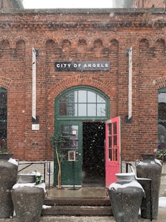 A brick building facade with a green arched door stands under a sign that reads 'City of Angels'. The door is ajar with a bright pink panel. Large decorative pots, some with snow on top, are placed on either side of the entrance. There are signs and banners hanging vertically next to the door, and light snow is gently falling.