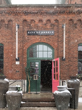 A brick building facade with a green arched door stands under a sign that reads 'City of Angels'. The door is ajar with a bright pink panel. Large decorative pots, some with snow on top, are placed on either side of the entrance. There are signs and banners hanging vertically next to the door, and light snow is gently falling.