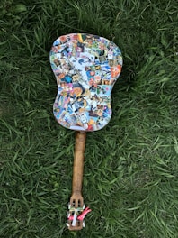 A guitar covered in colorful stickers lies on green grass. The body of the guitar features a variety of stickers, including cartoon characters and logos, while the neck has a wooden texture. The strings are not visible, and the mood is playful and eclectic.