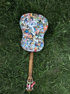 A guitar covered in colorful stickers lies on green grass. The body of the guitar features a variety of stickers, including cartoon characters and logos, while the neck has a wooden texture. The strings are not visible, and the mood is playful and eclectic.