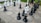 A large chess set with oversized black and white pieces placed on a checkered outdoor board made of stone tiles. A person is partially visible, interacting with the pieces. Surrounding the chessboard is a grassy area with a few scattered leaves.