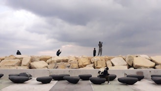 Several people are interacting with a coastal area that includes large, light-colored boulders and a series of smooth, rounded black objects. The sky is overcast, creating a dramatic atmosphere.