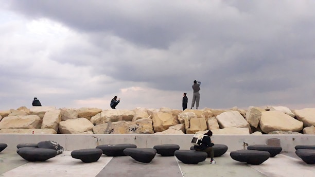 Several people are interacting with a coastal area that includes large, light-colored boulders and a series of smooth, rounded black objects. The sky is overcast, creating a dramatic atmosphere.