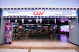 A brightly lit cinema entrance with a marquee displaying various messages. There is a large neon logo above the entrance. Inside, customers are seated at a ticket area, and there are large screens displaying movie-related content. Several decorations hang from the ceiling, adding a festive touch.