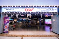 A brightly lit cinema entrance with a marquee displaying various messages. There is a large neon logo above the entrance. Inside, customers are seated at a ticket area, and there are large screens displaying movie-related content. Several decorations hang from the ceiling, adding a festive touch.