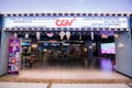 A brightly lit cinema entrance with a marquee displaying various messages. There is a large neon logo above the entrance. Inside, customers are seated at a ticket area, and there are large screens displaying movie-related content. Several decorations hang from the ceiling, adding a festive touch.