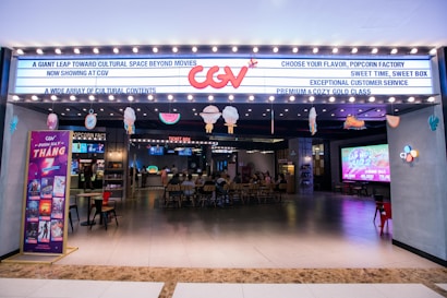 A brightly lit cinema entrance with a marquee displaying various messages. There is a large neon logo above the entrance. Inside, customers are seated at a ticket area, and there are large screens displaying movie-related content. Several decorations hang from the ceiling, adding a festive touch.