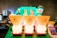 Three illuminated containers shaped like monster figures with one eye and comical expressions are arranged in a row on a counter. Each container has a drink within, topped with colorful lids featuring small character figures. The background includes a neon green sign with text, beverage machines, and promotional materials with vibrant colors.