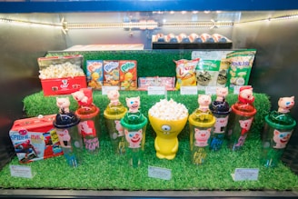 An assortment of themed products displayed on artificial grass, including popcorn tubs with cartoon decorations, pig-themed plastic cups with figurines on top, snack packets, and a Spider-Man-themed lunchbox filled with popcorn.