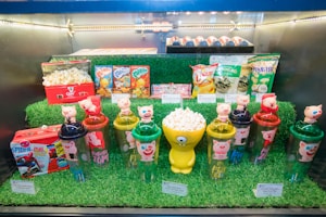 An assortment of themed products displayed on artificial grass, including popcorn tubs with cartoon decorations, pig-themed plastic cups with figurines on top, snack packets, and a Spider-Man-themed lunchbox filled with popcorn.