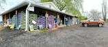 A warehouse with stacks of large bags, possibly of soil or gardening supplies, arranged on wooden pallets. The building has an overhanging roof with a sign reading 'Exit One Way'. A brown SUV is parked on a gravel surface near the entrance. There are trees with fresh green leaves in the background, suggesting a spring setting.