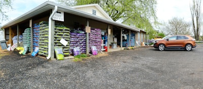 A warehouse with stacks of large bags, possibly of soil or gardening supplies, arranged on wooden pallets. The building has an overhanging roof with a sign reading 'Exit One Way'. A brown SUV is parked on a gravel surface near the entrance. There are trees with fresh green leaves in the background, suggesting a spring setting.