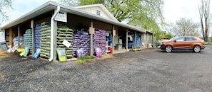 A warehouse with stacks of large bags, possibly of soil or gardening supplies, arranged on wooden pallets. The building has an overhanging roof with a sign reading 'Exit One Way'. A brown SUV is parked on a gravel surface near the entrance. There are trees with fresh green leaves in the background, suggesting a spring setting.