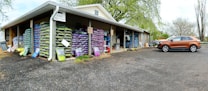 A warehouse with stacks of large bags, possibly of soil or gardening supplies, arranged on wooden pallets. The building has an overhanging roof with a sign reading 'Exit One Way'. A brown SUV is parked on a gravel surface near the entrance. There are trees with fresh green leaves in the background, suggesting a spring setting.