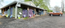 A warehouse with stacks of large bags, possibly of soil or gardening supplies, arranged on wooden pallets. The building has an overhanging roof with a sign reading 'Exit One Way'. A brown SUV is parked on a gravel surface near the entrance. There are trees with fresh green leaves in the background, suggesting a spring setting.