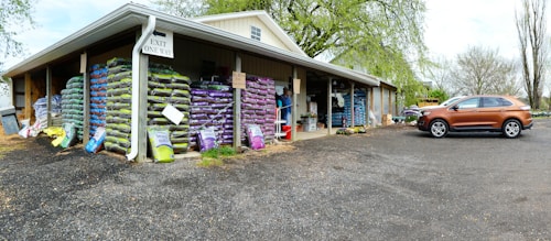 A warehouse with stacks of large bags, possibly of soil or gardening supplies, arranged on wooden pallets. The building has an overhanging roof with a sign reading 'Exit One Way'. A brown SUV is parked on a gravel surface near the entrance. There are trees with fresh green leaves in the background, suggesting a spring setting.