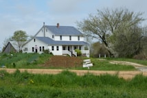 A rustic farmhouse with a white exterior and blue roof stands amidst lush greenery. A dirt path leads up to the house, surrounded by a vibrant garden with various plants. There is a sign in the front indicating 'Plants and Produce' for sale. A person wearing a hat is seen walking towards the house.