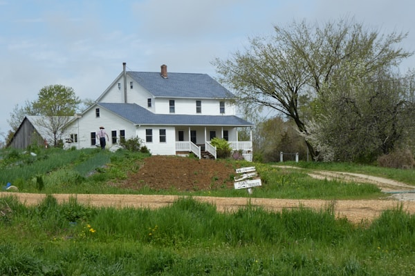 A rustic farmhouse with a white exterior and blue roof stands amidst lush greenery. A dirt path leads up to the house, surrounded by a vibrant garden with various plants. There is a sign in the front indicating 'Plants and Produce' for sale. A person wearing a hat is seen walking towards the house.