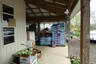 A covered outdoor area with stacks of gardening soil bags neatly arranged on wooden pallets. There are boxes of fresh vegetables, including cabbages, on the ground next to a wall. Various garden supplies and a hanging butterfly decoration are also visible. The area is supported by wooden posts and has a metal roof. In the background, trees and a portion of a white car can be seen.