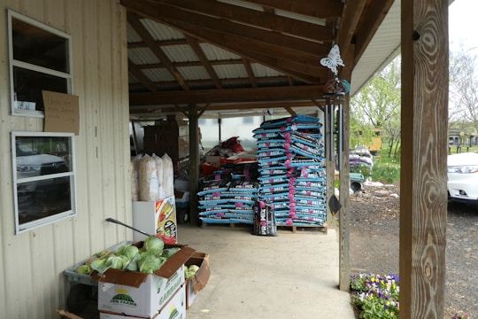 A covered outdoor area with stacks of gardening soil bags neatly arranged on wooden pallets. There are boxes of fresh vegetables, including cabbages, on the ground next to a wall. Various garden supplies and a hanging butterfly decoration are also visible. The area is supported by wooden posts and has a metal roof. In the background, trees and a portion of a white car can be seen.