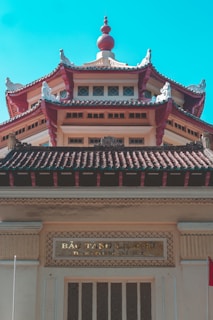 An intricately designed building features traditional Asian architectural elements, including curved roofs with ornate carvings and decorative details. The structure is adorned with a distinctive red and beige color scheme, highlighted by a central tower topped with an ornate finial. A sign with Vietnamese writing is visible above the entrance.