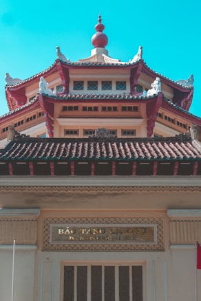 An intricately designed building features traditional Asian architectural elements, including curved roofs with ornate carvings and decorative details. The structure is adorned with a distinctive red and beige color scheme, highlighted by a central tower topped with an ornate finial. A sign with Vietnamese writing is visible above the entrance.