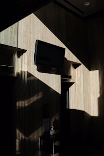 A dimly lit interior scene with a wall-mounted TV casting bold, geometric shadows. The wall has a textured, stone-like appearance, and there is a small table with a hand sanitizer bottle in the foreground. The contrast between the light and shadows creates a dramatic effect.