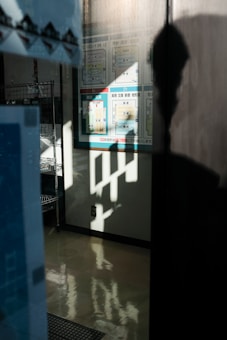 A partial reflection on a polished floor creating an abstract pattern. Wall signage and shelving units are visible in the background, with silhouette of a person partially visible.