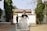 A boy is holding a cricket bat and standing outdoors. The setting appears to be a backyard or garden area with some trees and a gravel pathway. There is another person in the background, slightly out of focus, near a gate. A white plastic chair is placed in the foreground.