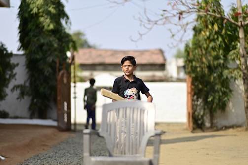 A boy is holding a cricket bat and standing outdoors. The setting appears to be a backyard or garden area with some trees and a gravel pathway. There is another person in the background, slightly out of focus, near a gate. A white plastic chair is placed in the foreground.