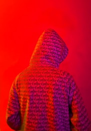 A person wearing a hooded sweatshirt with the words 'fear of god' printed repeatedly across the fabric. The background is a vivid red color, creating a striking contrast with the purple and orange hues of the hoodie.