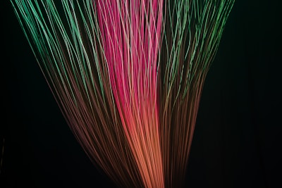 Multicolored light strands extend diagonally across a dark background, creating a dynamic and mesmerizing visual effect. The light streaks shift from green to pink and orange hues, giving the impression of motion and energy.