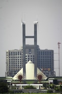 A tall, modern building with twin spires topped by wind turbines stands prominently in front of a large, unfinished structure. The building features geometric designs and is flanked by two star-shaped sculptures. A crane is visible in the background, highlighting ongoing construction.