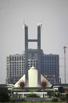 A tall, modern building with twin spires topped by wind turbines stands prominently in front of a large, unfinished structure. The building features geometric designs and is flanked by two star-shaped sculptures. A crane is visible in the background, highlighting ongoing construction.