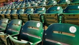 A close-up view of several rows of green stadium seats, each labeled with sequential numbers. The seats are empty, and in the background, a few people can be seen sitting further back.